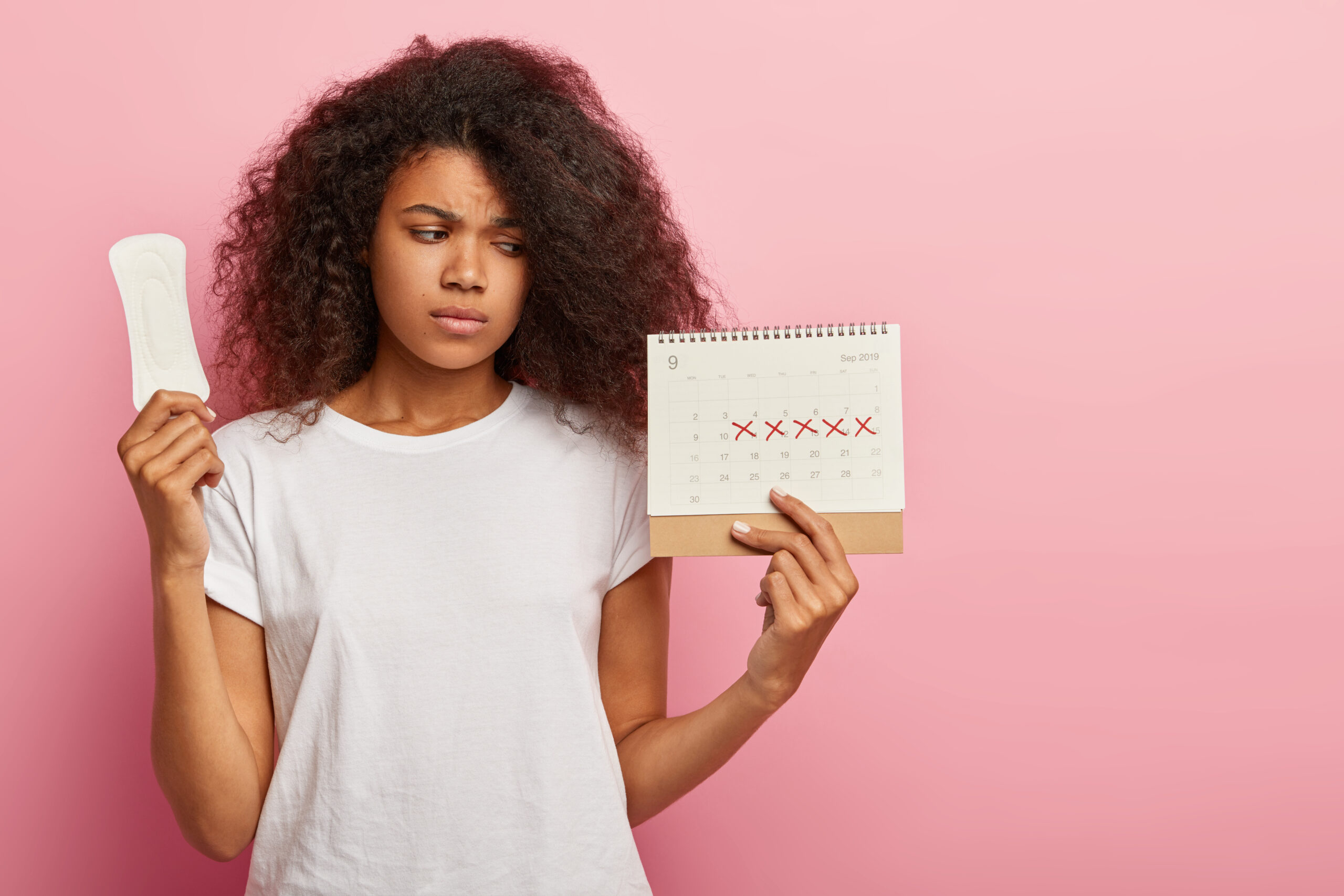 Photo of discontent lovely curly haired woman looks at calendar with marked pms days, holds sanitary napkin for hygiene protection, has bad mood during menstruation, wears casual white t shirt