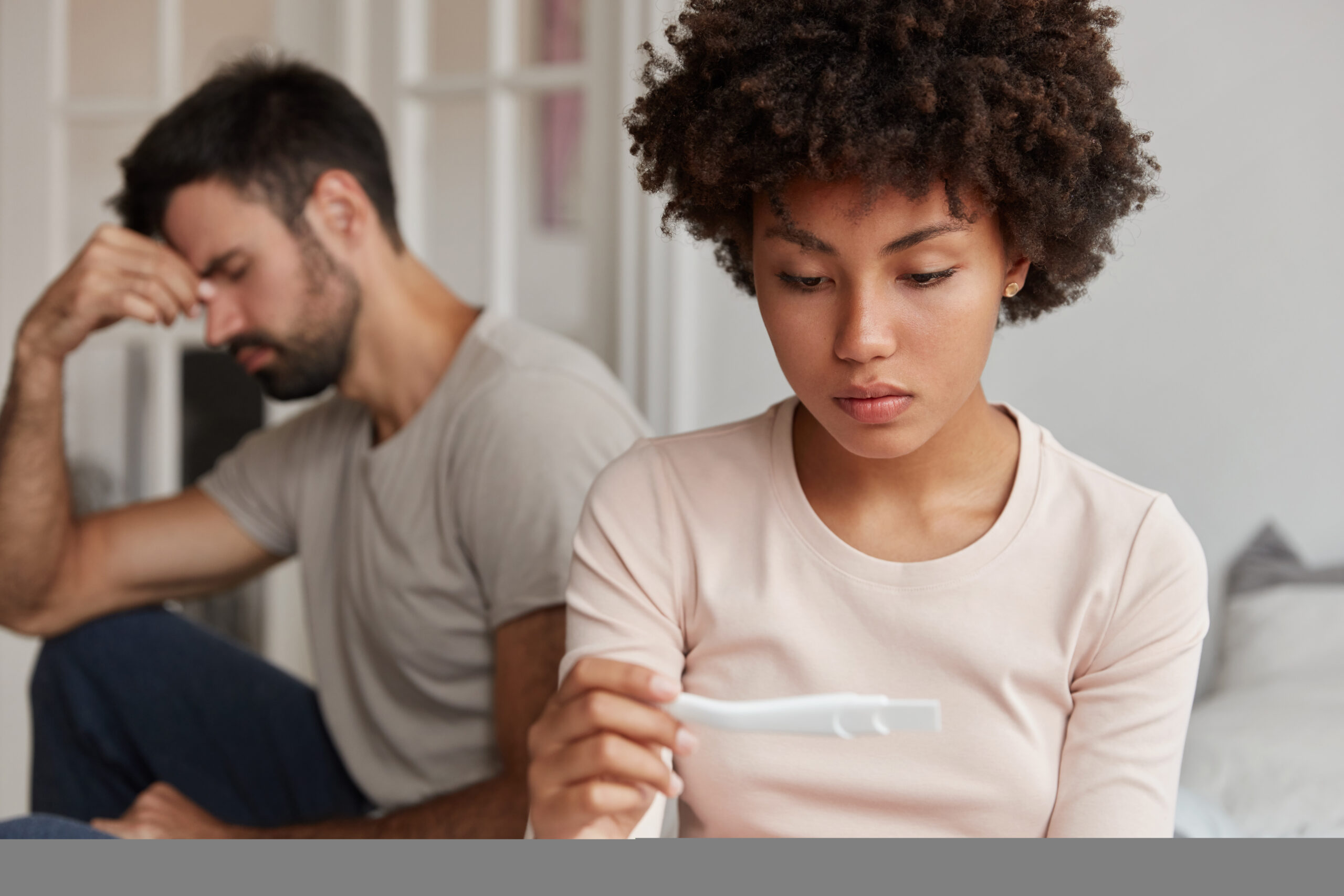 Upset dark skinned attractive woman looks at pregnancy test, suffers from infertility, frustrated husband sits near at edge of bed. Family couple have problem, cant have children for long time