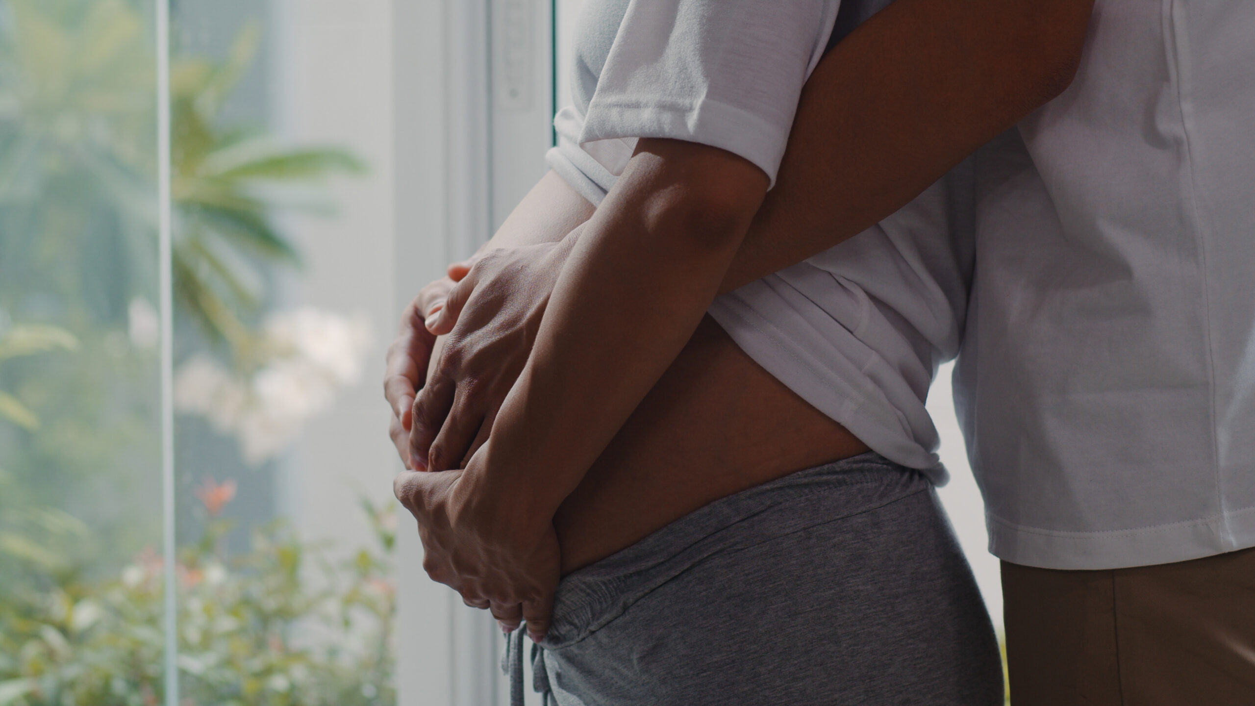 Young Asian Pregnant couple hug and holding belly talking with their child. Mom and Dad feeling happy smiling peaceful while take care baby, pregnancy near window in living room at home concept.