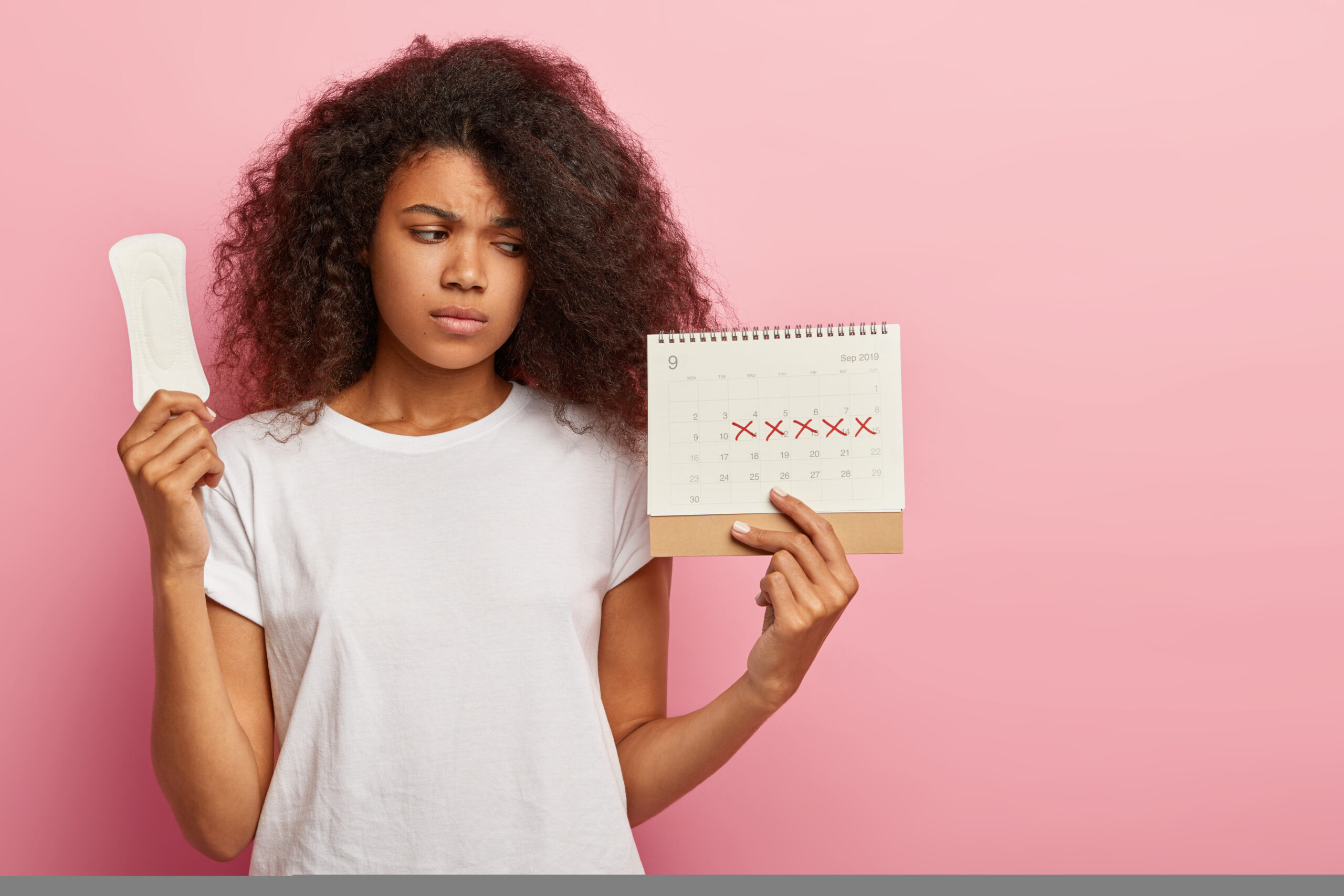 Photo of discontent lovely curly haired woman looks at calendar with marked pms days, holds sanitary napkin for hygiene protection, has bad mood during menstruation, wears casual white t shirt